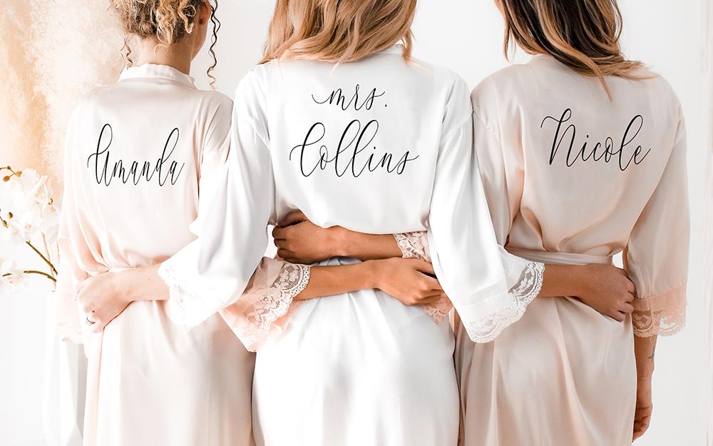 Three ladies stand with their back facing the viewer interlocking arms wearing delicate satin lace bridesmaid robes that have their names in dainty cursive on the back.
