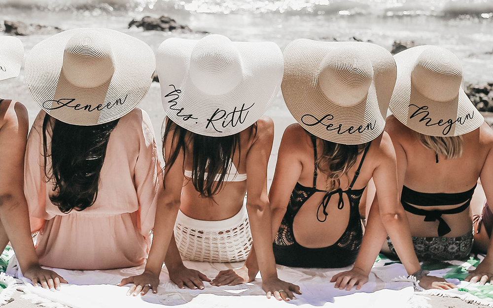 Five bridesmaids are sitting on the beach in the sand side by side with their back facing the viewer showing off wide brimmed sunhats that feature their names in black dainty cursive.