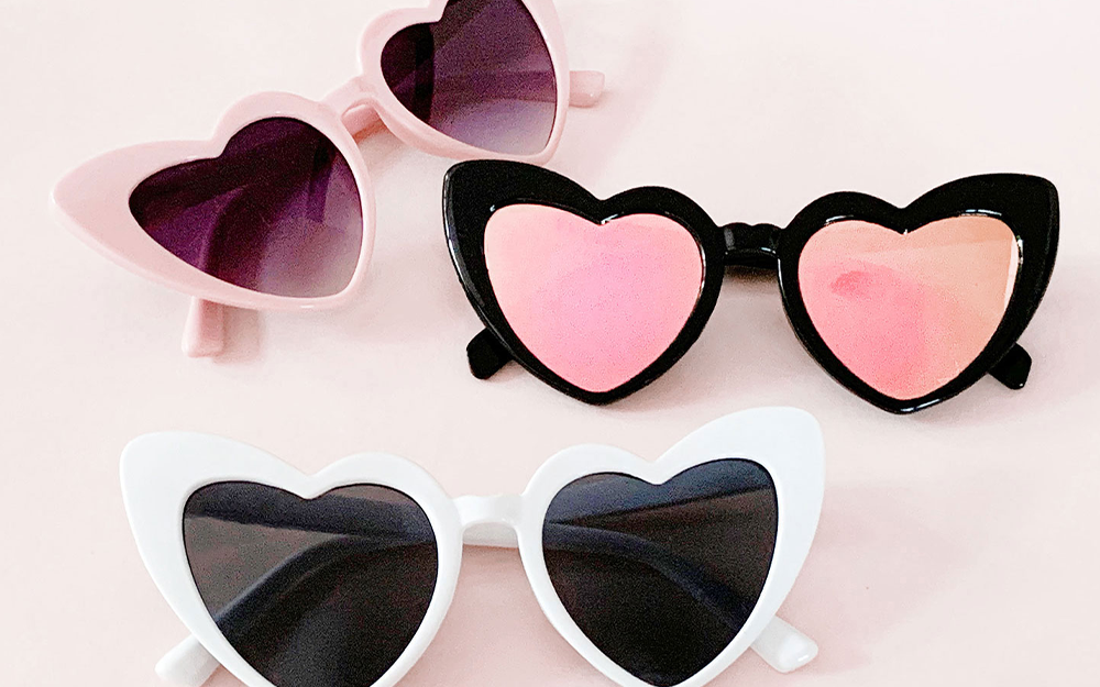 Three heart shaped sunglasses in white, pink, and black are resting on a baby pink background.
