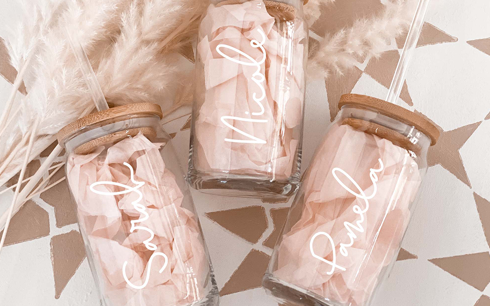 Three clear glass can style coffee cups are laying on the ground with eco-friendly sealed bamboo lids and straw featuring your bridesmaid names in white cursive text.