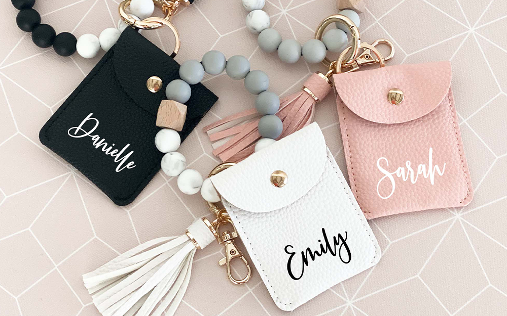 Three small wallets that are black, pink, and white are laying facing up showcasing her name in black or white cursive text. Wristlet wallets have a bulk beaded wrist bracelet.