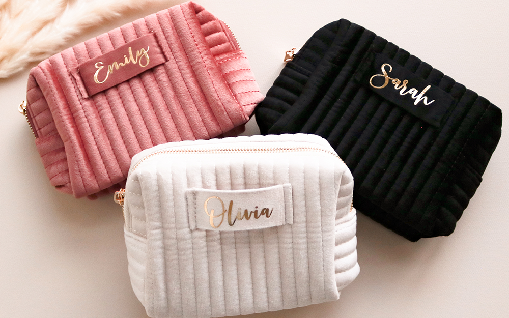 Three plush velvet cosmetic bags in mauve, white, and black laying on a light beige background featuring shimmery gold cursive text in her name.
